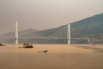 Fengdu, Chongqing, China - May 8, 2010: Yangtze River. Wide landscape of H-shaped pylons of suspension bridge over brown water with boat traffice under smog sky with green hills on shore,