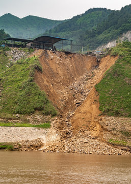Huangqikou, Chongqing, China - May 8, 2010: Yangtze River. Collapsed shoreliine with brown dirt landslide under green forested mountains behind brown water. Blue industrial building.