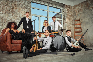 Obraz na płótnie Canvas multi ethnic jazz band posing on a leather sofa against the window in loft. Bass guitar player, electric guitar player, saxophonist and drummer at loft. Jazz music and jam session concept. passion for