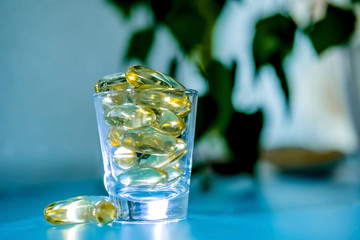 A glass cup with omega 3 vitamins stands on a blue table near the window, near a birch tree,.