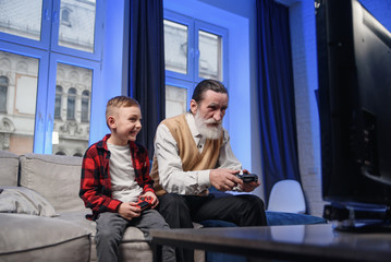 Grandfather and grandson are playing exciting video games on computer with gamepads at home.