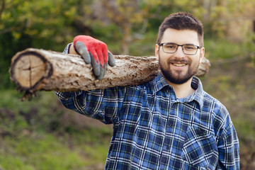 Portrait of young adult caucasian man male worker woodcutter wearing shirt beard and protective working gloves holding a log tree trunk on shoulder in yard outdoor in day smiling front view