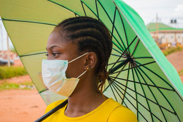 young beautiful african lady wearing face mask to prevent, preventing, prevented herself from the outbreak in the society and holding a umbrella