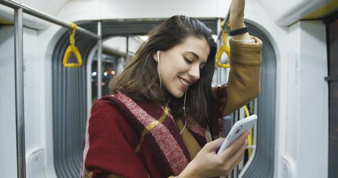 Caucasian young beautiful woman in coat, scarf and headphones listening to music on smartphone and coming back home at night. Attractive stylish female passanger standing in tram and holding phone.
