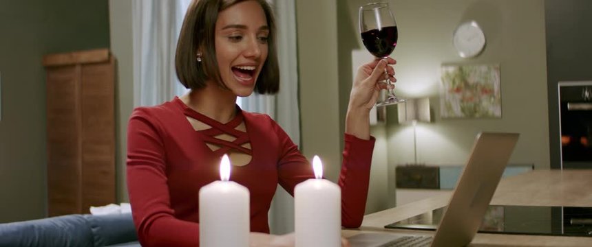 Beautiful woman in a red dress having a glass of wine chatting, online date with partner. Stay home, quarantine life. Shot with anamorphic lens