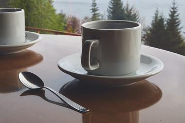 Taza de café sobre mesa de madera, con vista a lago, bosque y montañas.