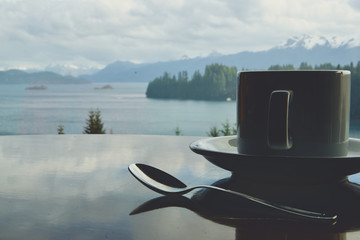 Taza de café sobre mesa de madera, con vista a lago, bosque y montañas.