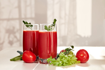 Two glasses with fresh tomato juice and ripe tomatoes on white table near window.