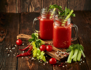 Two glasses with fresh tomato juice, celery, parsley and ripe tomatoes on dark brown wooden background.