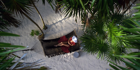 Woman rests in loose red summer dress and hat hanging in hammock surrounded by palm trees at sunset - obrazy, fototapety, plakaty