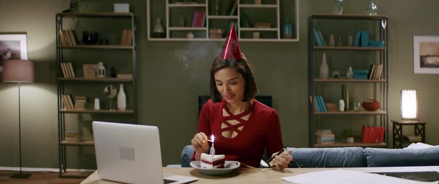 Beautiful woman in a party cap lights a candle on a cake, having an online birthday party with friends. Stay home, quarantine life. Shot with anamorphic lens
