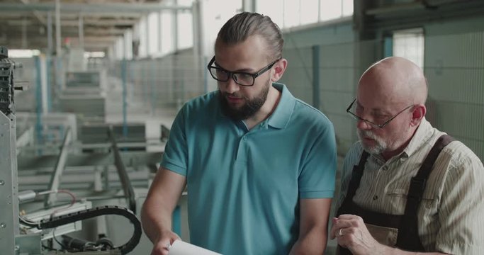 Two engineers stand at factory facility, have discussion and use materials notes