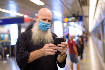 Mature bald bearded man with mask using phone at the subway train station