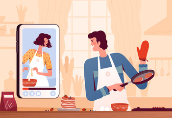 A young man watches a video recipe on the phone and prepares pancakes at home in the kitchen. The concept of online recipes and home cooking