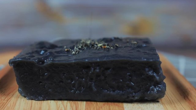 Sprinkled white coconuts and black sasame and sugar on top of dark grey Rhombus Thai dessert (Thai called name is Kanom paek poon) in a wooden plate , rotating on a wooden plate