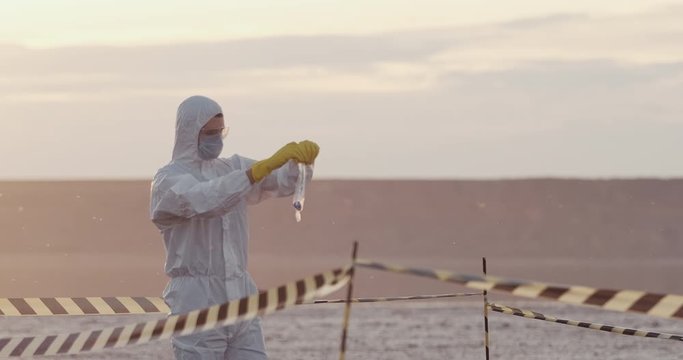 A scientist in a protective suit puts a ground sample contaminated with biological substances in a sealed bag, his partner takes pictures of the sample.