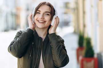 Adorable lady in standing in city and listening favorite song in headphones. Outdoor portrait of dreamy european girl wears white earphones.