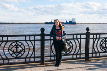 A white woman of forty years stands happy on the embankment, substituting her face and hair in the wind, against the background of the river with freight river transport and blue cloudy sky. 
