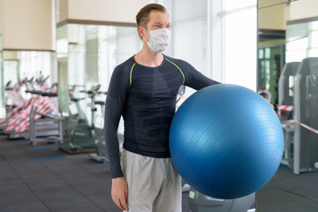Young man with mask thinking while holding exercise ball at gym during corona virus covid-19