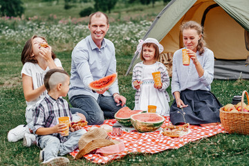 Happy family eating watermelon at picnic in meadow near the tent. Family Enjoying Camping Holiday In Countryside