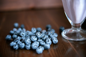 a pile of blueberries on a wooden table next to a clear milkshake bowl close up