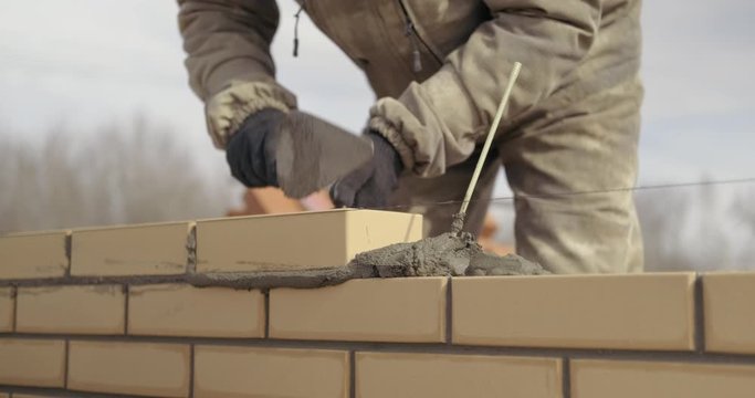 Bricklayer works at construction site, lays bricks in masonry, builds house, erects brick wall, yellow cladding brick, works as trowel, open air, in special uniform, professional work, smooth seams