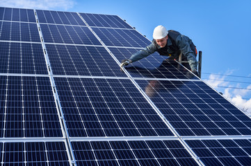 Electrician standing on ladder and installing blue photovoltaic solar panel system. Man technician in safety helmet under blue sky. Concept of alternative energy and power sustainable resources.