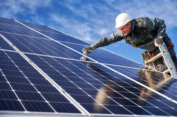 Side view of joyful male technician in safety helmet standing on ladder and installing photovoltaic...