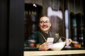 Young smiling girl sitting in restaurant near window, eating her healthy lunch, holding smart phone and looking away. Picture taken from outside.