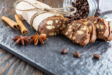 Chocolate salami on a stone slab, cinnamon and star anise sticks lie nearby, coffee beans pour out from a glass jar, shallow depth of field, selective focus. Breakfast dessert concept