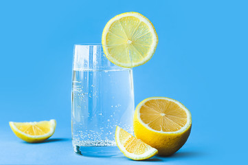 translucent glass of water with gases and bubbles, with a round slice of lemon on a glass and a whole lemon on blue background. Refreshment in the heat. Healthy lifestyle and nutrition