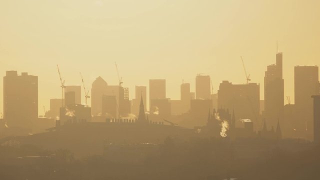 Sunrise in hot weather over London, UK. Pollution, smog and smoke from chimneys are seen in the foreground and the hazy outline of skyscrapers on the skyline are seen in the background.