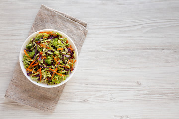 Homemade Raw Shredded Broccoli Slaw in a white bowl on a white wooden surface, top view. Overhead, from above, flat lay. Copy space.