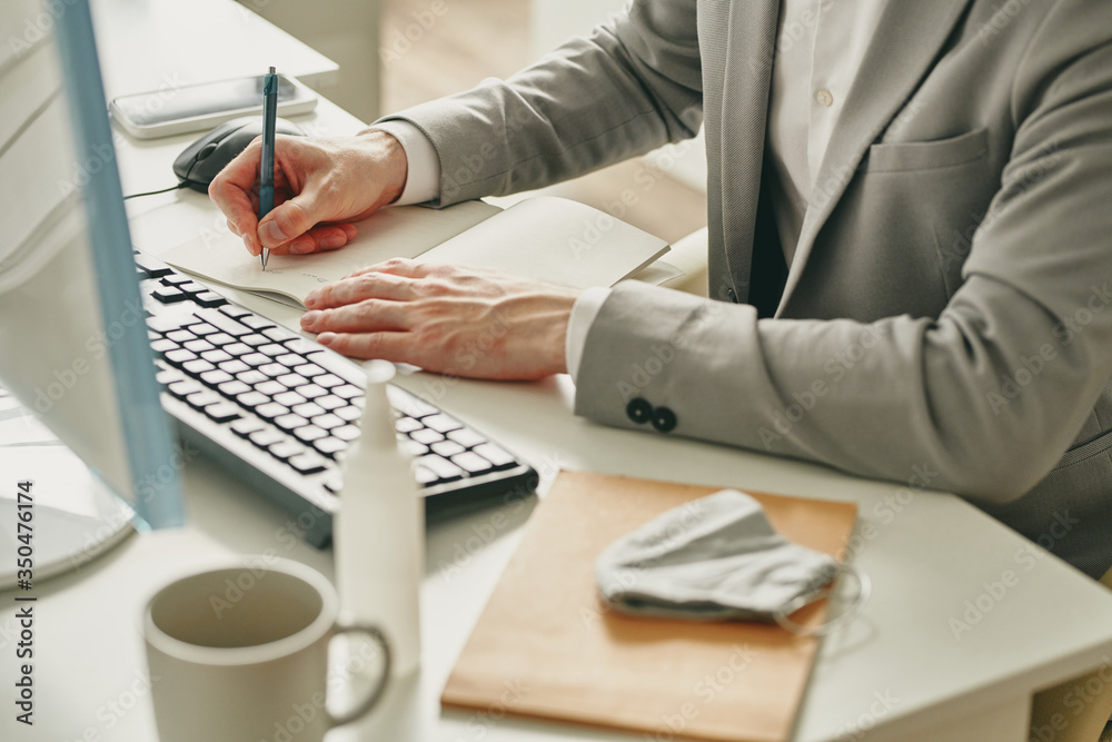 Poster Close-up of businessman sitting at desk with sanitizer and mask and making notes in notepad while working in office during coronavirus quarantine