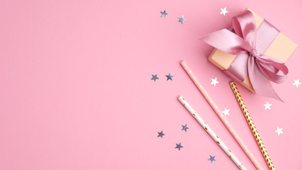 Gift box and drinking straws with confetti stars on pink table top view. Girls birthday, wedding or Christmas party concept.