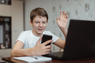 Happy boy using laptop computer and phone to communicate with friends on the Internet at home and waving hand. Online video chat. Social distance during quarantine