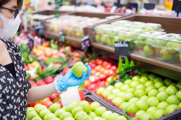 Asian woman wearing nitrile gloves and holding green apple fruit in supermarket or grocery, protect coronavirus inflection. Hygiene, new normal and life after covid-19 pandemic