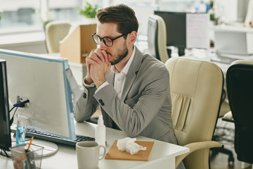 Nervous young businessman in eyeglasses sitting with closed eyes and trying to focus on work in empty office during coronavirus