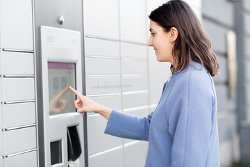 Fototapeta na wymiar mail delivery and post service concept - happy smiling woman choosing operation on outdoor automated parcel machine's touch screen