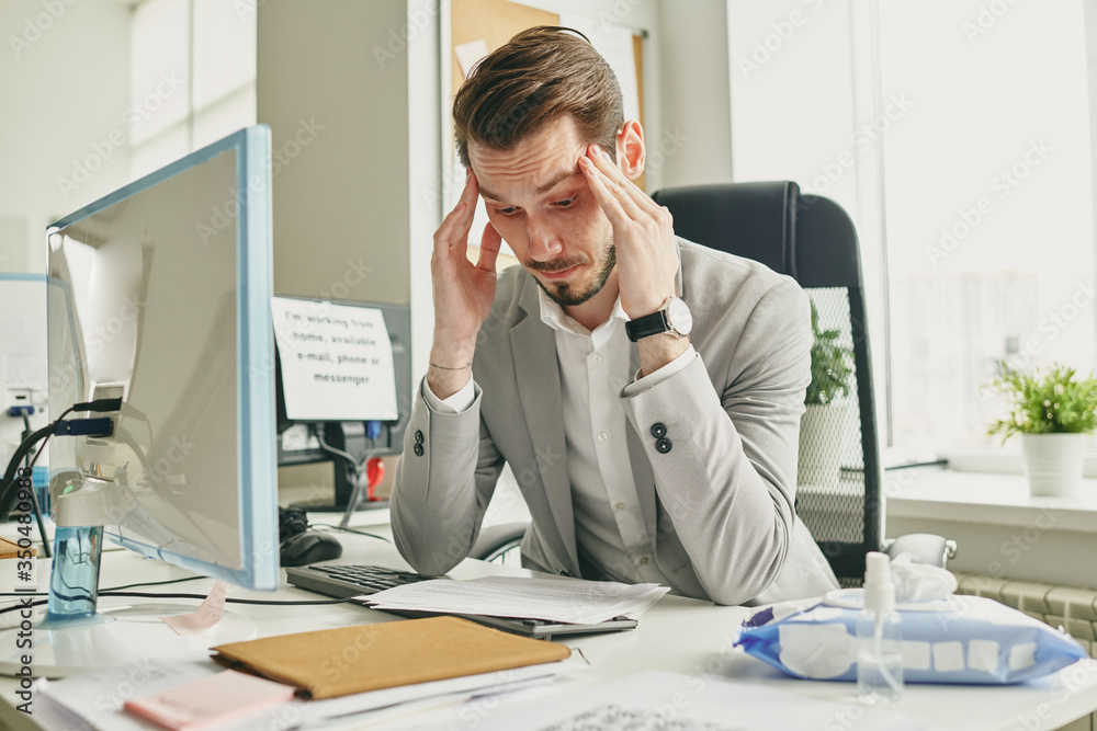 Poster Confused young businessman in jacket sitting at desk in empty office and massaging temples while reading agreement