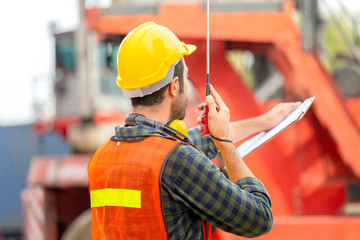 Foreman in hardhat and safety vest holding holding clipboard checklist and talks on two-way radio control loading containers box from cargo