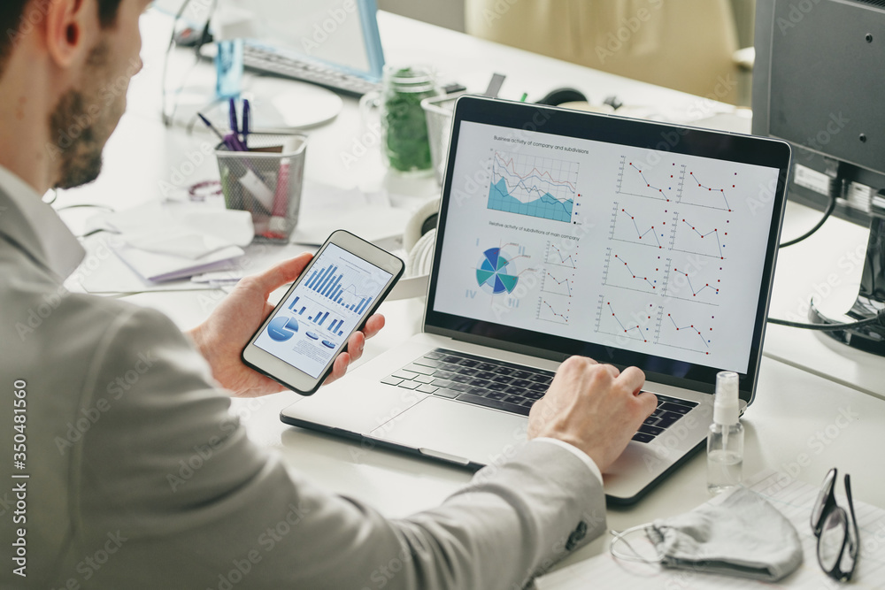 Aufkleber Over shoulder view of businessman using smartphone and laptop while analyzing sales graphs during quarantine