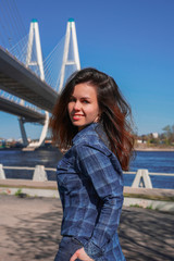 A brunette girl with long hair in a blue shirt stands against the background of a cable stayed bridge on the embankment