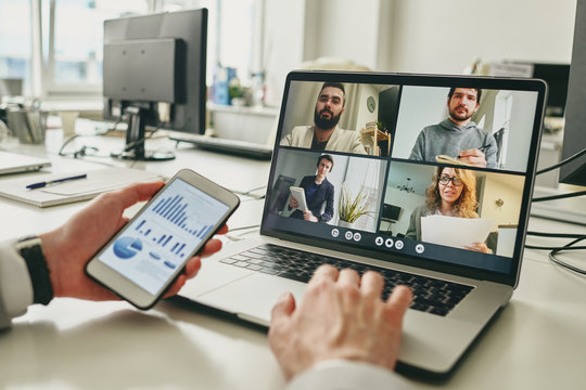 Close-up of businessman working in empty office and using smartphone to view graph while discussing sales with colleagues via video conferencing app