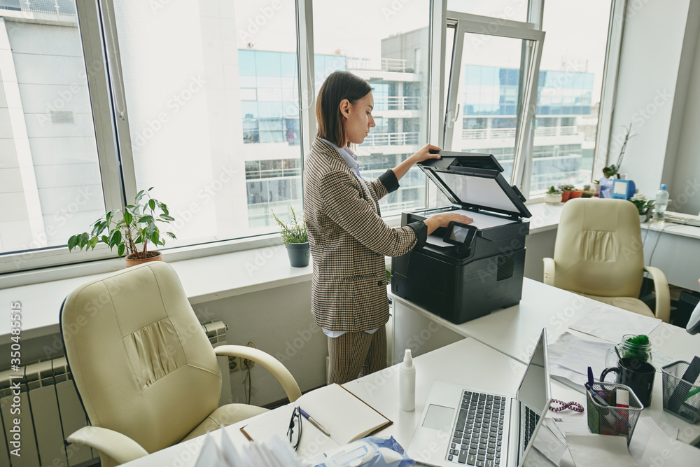 Poster young female manager in jacket standing at scanner and making copy of document in empty office durin