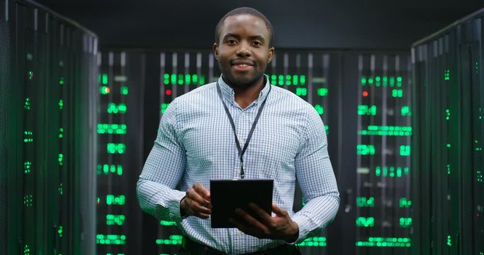 Portrait of young handsome African American male big data specialist wih tablet device in hands smiling to camera in servers room. Happy man in database center. Digital storage concept.