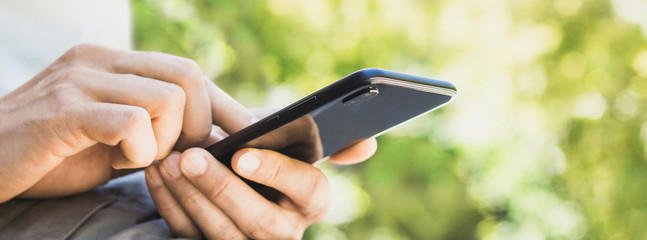 Young man texting on his smartphone in the city. Close up of adult male hand using mobile phone outdoor, panoramic banner
