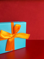 Bright blue gift box with an orange ribbon on a contrasting burgundy background. Happy birthday greeting card. Closed box with a gift on a red background. Copy space for text.