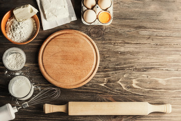 Baking cooking ingredients flour, eggs, rolling pin, butter, cottage cheese and a wooden round board on a wooden background. View from above. Copy space. The composition of the test
