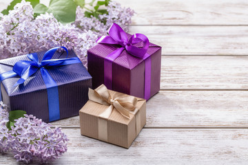 Gifts in holiday boxes with ribbons and bows. Branches of flowers of lilac. Wooden background, studio shot.